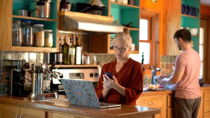 Teen son, blurry in the distance washes dishes as mature mother uses phone and works on laptop computer in kitchen.
