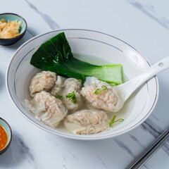 A delicate bowl of wonton soup with bok choy, served with fresh green onions, fried garlic, and spicy chili sauce on a white marble table, complemented by chopsticks