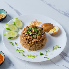 A white plate of Thai-style rice topped with minced chicken, served with sliced cucumber, a halved boiled egg, green onions, fried garlic, and chili sauce, alongside a soup bowl,