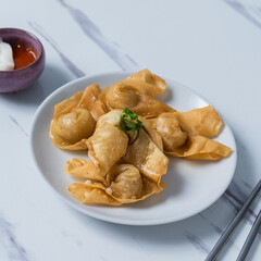 A white plate of crispy fried wontons garnished with green onions, served a small bowl of sweet chili sauce and mayonnaise on a marble surface with chopsticks, perfect for Asian appetizer content.