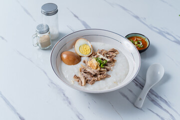 A white bowl of congee topped with shredded chicken, a halved boiled egg, and fried garlic, served with chili sauce and condiments on a marble surface, ideal for Asian breakfast content