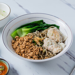 A white bowl of Asian noodles topped with minced pork, bok choy, and wontons, served with a bowl of...