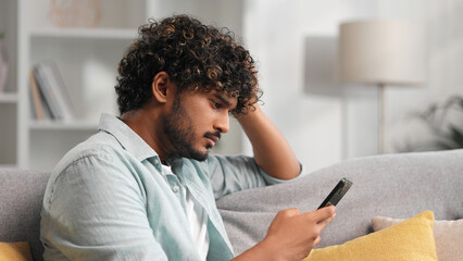 Worried young Indian boy sitting on sofa looking at her phone and does not understand why it is not working. The angry student in glasses does not believe the bad news he saw.