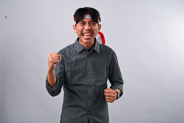 Indonesian man celebrating with fists raised, wearing a grey shirt and a red-and-white headband. He...