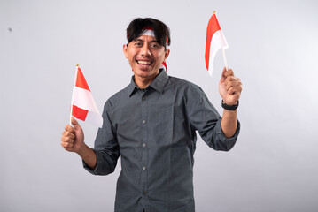 A cheerful Indonesian man wearing a gray shirt and headband, holding two Indonesian flags. He is...