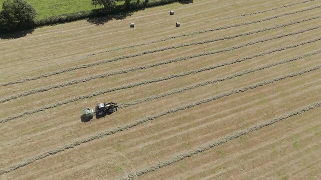 Drone aerial shot of a tractor pulling a baler through a field, collecting freshly cut grass and forming it into neat bales, the scene set against a picturesque rural backdrop