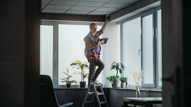 Male mature technician stands on ladder, installs CCTV camera in office room using tablet computer with professional software. Monitoring and tracking. Security system, video surveillance and privacy.