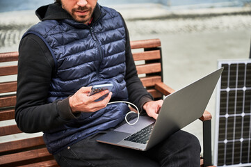 Cropped view of man sits on wooden bench, working on laptop and smartphone, both connected to solar...