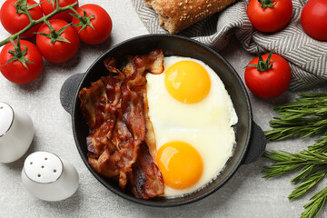 Tasty bacon and eggs in dish among products on gray textured table, flat lay