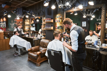 Professional barber using comb and electric shaver to cut side hair. Male customer getting stylish haircut. Side view of man on background of other hairdresser.