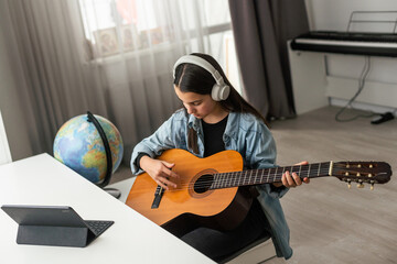 Latin girl with headphones listening to her online music lessons. Artistic kid playing the acoustic guitar and learning the chords