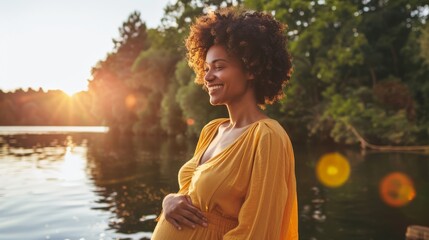A joyful pregnant woman stands by the lakeside at sunset in a flowing yellow dress, gently cradling her belly, basking in the warm glow and natural beauty of the setting.
