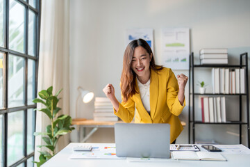 Asian businesswoman celebrating success raising fists looking at laptop at office