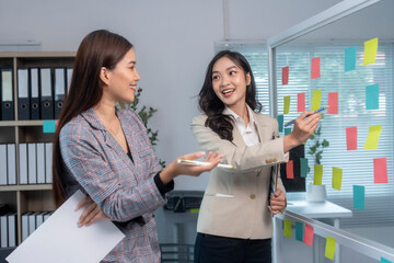 Two businesswomen discussing and brainstorming using sticky notes on glass wall in office