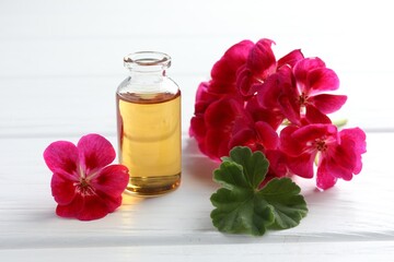 Bottle of geranium essential oil and beautiful flowers on white wooden table, closeup