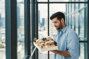 Businessman in casual outfit holding house model while checking house construction. Architect engineer inspect building model while standing near window with skyscraper. Civil engineering. Tracery.