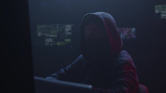 A mysterious shadowy figure clad in a hoodie, intently focused on multiple computer screens in a dark environment