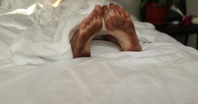 A man with very dirty feet wakes up in bed with clean white linen. Funny situation. High quality 4k footage