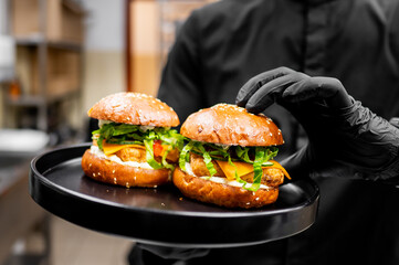 A close-up of two gourmet burgers with fresh toppings on a tray held by a person in black attire and gloves, showcasing delicious fast food. Perfect for culinary content or advertising.