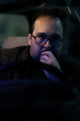 A forty-year-old man in winter clothes and glasses rides in a retro car. He looks thoughtful and tired.