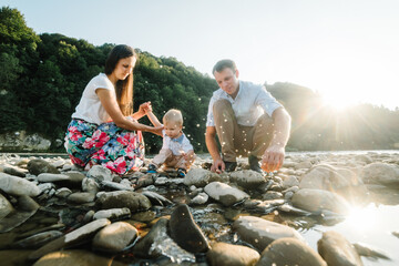 Mother, father, baby son sitting and playing on beach near lake. Happy family holiday on summer day. Spending time together in mountains. Parents and kid throw stones into the water at sunset.