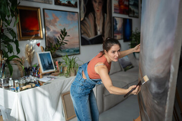 Woman painter crafts landscape on canvas of easel in loft studio. Talented serious female artist...