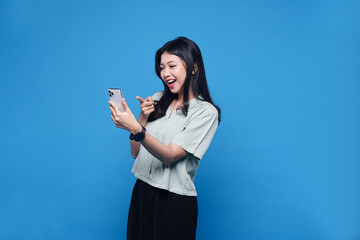 Happy Asian young woman using mobile smartphone, read good news online, laugh excited getting offer, great positive surprise, celebrate success on smartphone. Smile girl isolated on blue background