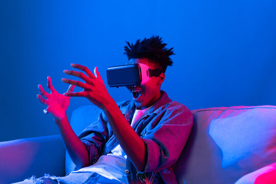 Young African American looking VR enjoy watching fantastic 3D pointing interesting object technology blue neon light virtual reality metaverse new world at living room sitting on sofa. Contrivance.