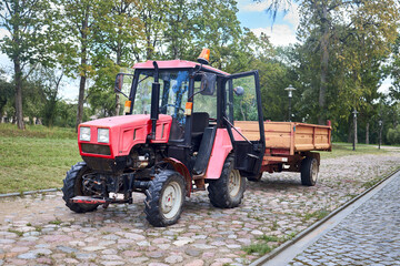 Small mini red tractor with trailer standing at stone farm road sunny summer day. Small agricultural machinery. Rural country farmland background.