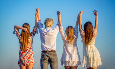 Fototapeta premium Young friends holding hands while standing back at camera outdoors. Back view. Company of young girls and guys are standing in the field on a summer day and holding their hands up