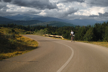 Adventure cyclist rides a gravel bike along a rugged, scenic gravel road. The backdrop features rolling hills and a cloudy sky, capturing the essence of adventure.