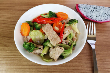 Stir-fried pork, broccoli, carrots, chili in a porcelain plate. Place a fork on the side. and sliced ​​dragon fruit on a wooden table