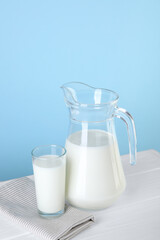 Jug and glass of fresh milk on white wooden table against light blue background