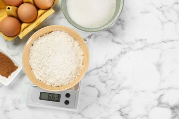 Flat lay composition of kitchen scale with bowl of flour and products on white marble table. Space for text