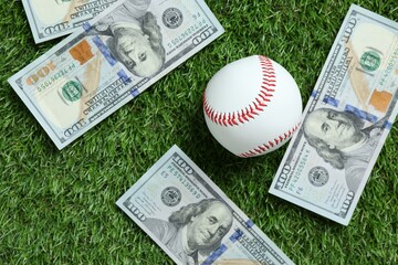 Baseball ball and dollar banknotes on green grass outdoors, flat lay
