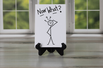 Sign with stick figure scratching head with question marks, Now What? Hand written.