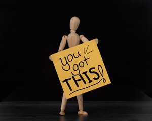 You Got This! Handwritten on yellow sign. Held by wooden mannequin.