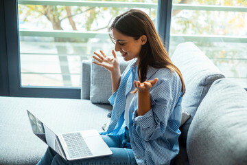 Young woman sitting on a sofa at home, smiling and having a joyful conversation with friends or a partner on her computer. Gesticulating and enjoying a fun moment, seen from the side, in a cozy relax