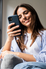 Young, modern woman in casual clothes smiles while using her smartphone at home, scrolling social...