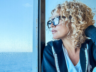 Portrait of a beautiful middle-aged woman with blonde curly hair and glasses, thoughtfully gazing with a dreamy expression against a deep blue ocean background, contemplating life and travel
