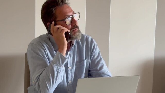 Busy mature man office worker manager consulting client by telephone checking information on laptop screen. Professional teleworker broker salesman offer goods services remotely using phone laptop