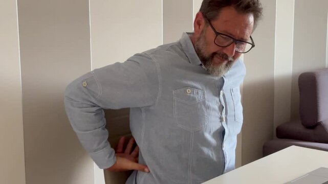 Senior man experiencing back pain while sitting at a desk in his home office, massaging his lower back due to age-related discomfort and poor posture from prolonged remote work in an uncomfortable cha