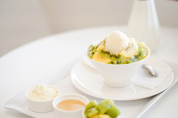 Refreshing shaved ice dessert served in a white bowl topped with a scoop of vanilla ice cream, with additional toppings and sauce on the side.