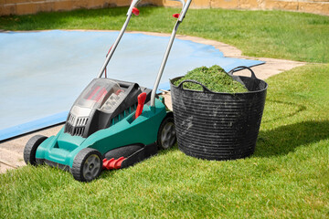 Electric lawn mower with rechargeable battery next to a work basket filled with the cut grass...