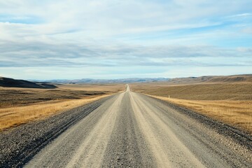 Fototapeta premium A lonely gravel road stretches out towards the distant horizon, winding its way through a barren, expansive landscape that lies beneath a dramatic, cloudy sky filled with muted light and shadows