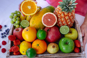 Wooden basket on white table full of fresh tropical colorful fruits. Female hands holding half orange fruit. Healthy eating and lifestyle