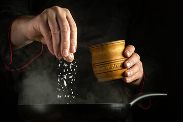 A cook adds salt to a hot frying pan with food being cooked by hand. The concept of cooking a dish in the kitchen with the hands of a chef.