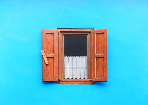 One isolated rustic window set in a blue facade, with open wooden shutters and a white lace curtain inside