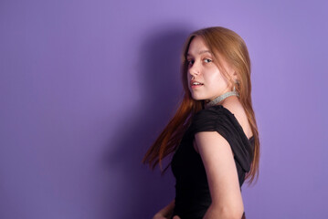 Fototapeta premium Young teenage girl trying on her new dress and new earrings and necklace on purple background, girl going on a date. The girl has beautiful red long hair