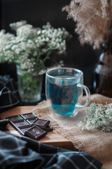 Blue cold beverage in transparent glass mug with chocolate on gunny and wooden tray, white flowers around, dark rustic lifestyle background in low key, idea of delicious dessert in the summer morning